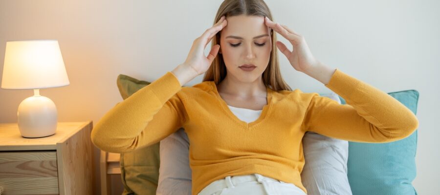 Young woman sitting in her bed and clutching her head in pain from overnight jaw clenching.